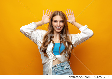 Young woman standing over yellow background doing funny gesture with hands over head as horns 94251263