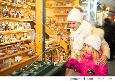 Little girl with happy mom buying figures and workpiece for creating Christmas scenes 94252456