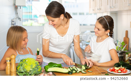 Cheerful woman with two little daughters prepare vegetable salad in the kitchen Cheerful woman with two little daughters prepare vegetable salad in the kitchen 94252660