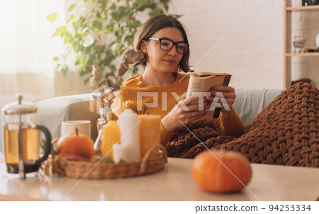 Lady reads a book in blanket on the sofa in front of teapot and candles 94253334