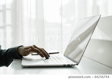 Man's hands using laptop computer with smartphone on white table at home Man's hands using laptop computer with smartphone on white table at home 94256744