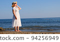 woman in a straw hat and white dress walks along the seashore and admires the blue sea on a sunny 94256949