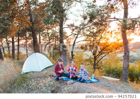 A cheerful company of two girls and a boy on a picnic in the middle of the forest. Children fry sausages on the fire, eat buns and have fun in nature. The concept of active recreation in the summer 94257021