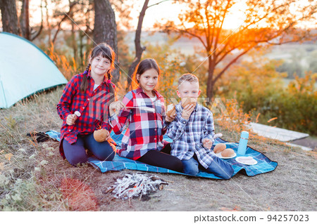 A cheerful company of two girls and a boy on a picnic in the middle of the forest. Children fry sausages on the fire, eat buns and have fun in nature. The concept of active recreation in the summer 94257023