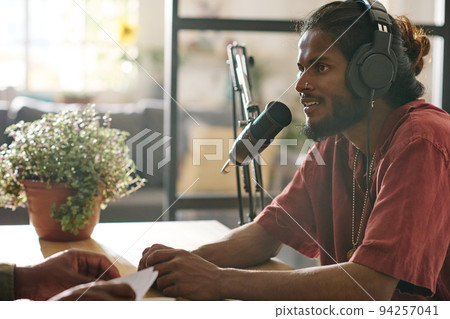 Side view of young multi-ethnic man sitting by desk in front of microphone Side view of young multi-ethnic man sitting by desk in front of microphone 94257041