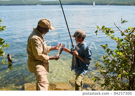 Side view of happy mature black man in activewear showing fish to his grandson Side view of happy mature black man in activewear showing fish to his grandson 94257300