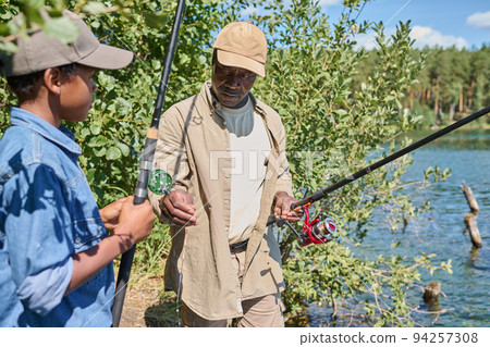 Aged African American man in casualwear holding rod while fishing with grandson Aged African American man in casualwear holding rod while fishing with grandson 94257308