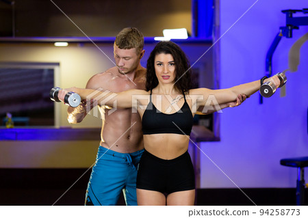 Young sports fitness trainer man trains young beautiful woman athlete in gym, shows exercises with dumbbells on biceps, legs 94258773