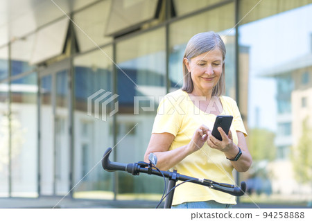 Portrait of a beautiful senior woman. Stands with a yellow bicycle near the office center, uses the phone. Gets a message. He's calling. He smiles. Active lifestyle Portrait of a beautiful senior woman. Stands with a yellow bicycle near the office center, uses the phone. Gets a message. He's calling. He smiles. Active lifestyle 94258888