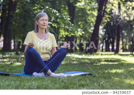 Senior gray-haired retired woman in the park meditates and performs breathing exercises, summer day in lotus position Senior gray-haired retired woman in the park meditates and performs breathing exercises, summer day in lotus position 94259037