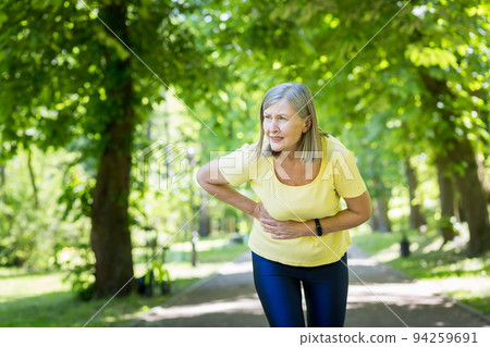 Senior beautiful woman running in the park, playing sports. Her stomach began to ache, she held on to her stomach, bent over in pain. Senior beautiful woman running in the park, playing sports. Her stomach began to ache, she held on to her stomach, bent over in pain. 94259691