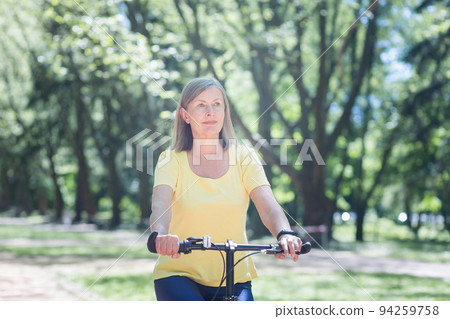 Active retired woman rides a bicycle in a summer park, a senior gray-haired retired woman walks 94259758