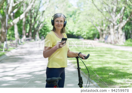 Senior beautiful woman walks in the park, rides a bike, listens to music on headphones, holds a phone. He stands with a bicycle in his hands, looks at the camera, smiles. Senior beautiful woman walks in the park, rides a bike, listens to music on headphones, holds a phone. He stands with a bicycle in his hands, looks at the camera, smiles. 94259978