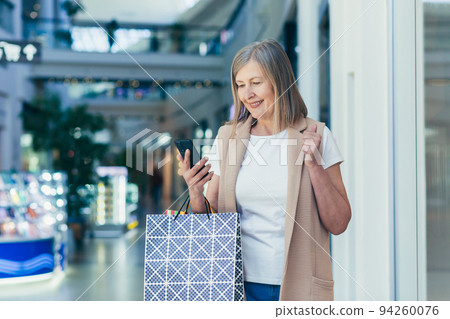 Portrait of a beautiful senior woman shopping in the mall. She uses the phone, makes purchases online, won a gift, rejoices. Holds paper shopping bags in her hands. She smiles, happy with her shopping 94260076