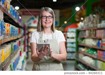 Portrait of senior woman in glasses consultant, employee in grocery store, supermarket. Stands with a tablet, accepts the goods, checks. In work clothes Portrait of senior woman in glasses consultant, employee in grocery store, supermarket. Stands with a tablet, accepts the goods, checks. In work clothes 94260311