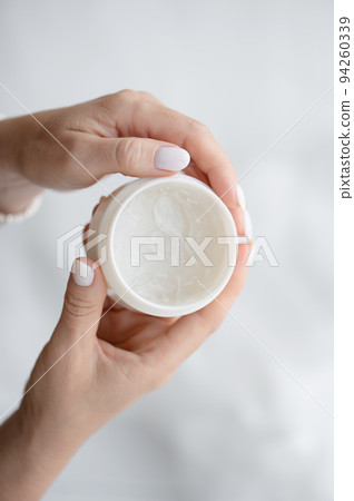 A woman's hands hold a white jar and use moisturizer. Taking care of her nails and clean, soft, smooth skin on her body. Front view. Close-up. A woman's hands hold a white jar and use moisturizer. Taking care of her nails and clean, soft, smooth skin on her body. Front view. Close-up. 94260339
