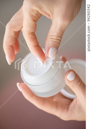 A woman's hands hold a white jar and use moisturizer. Taking care of her nails and clean, soft, smooth skin on her body. Front view. Close-up. 94260342