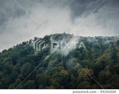 Peaceful fall scene with foggy clouds moving through the mixed forest on the top of a hill in a gloomy day. Natural autumn landscape in the woods, rainy weather with mist above the trees 94260880