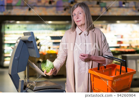 Self-service in the store. Portrait of a beautiful senior woman in a supermarket, shop. Weighs on the scales in the store cauliflower, vegetables, wants to buy. Holds a food basket in his hand Self-service in the store. Portrait of a beautiful senior woman in a supermarket, shop. Weighs on the scales in the store cauliflower, vegetables, wants to buy. Holds a food basket in his hand 94260904