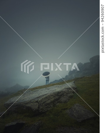Rear view of a lonely man with umbrella stands on a rocky hill covered by haze. Moody and emotional scene with a lone stranger silhouette under the rain Rear view of a lonely man with umbrella stands on a rocky hill covered by haze. Moody and emotional scene with a lone stranger silhouette under the rain 94260907