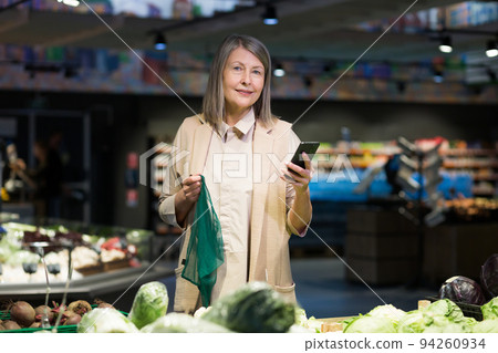 Senior woman in a supermarket chooses goods, looks at the electronic list on the phone 94260934