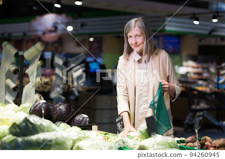 Portrait of a senior woman in a grocery supermarket, digs vegetables and puts in an ecological bag smiling and looking at the camera Portrait of a senior woman in a grocery supermarket, digs vegetables and puts in an ecological bag smiling and looking at the camera 94260945