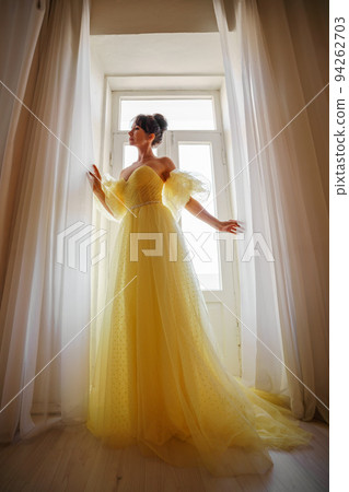 A woman's silhouette in a golden luxurious dress against the background of a window holds a curtain with her hands. Elegant lady in a yellow long silk dress with bare back, back view. A woman's silhouette in a golden luxurious dress against the background of a window holds a curtain with her hands. Elegant lady in a yellow long silk dress with bare back, back view. 94262703