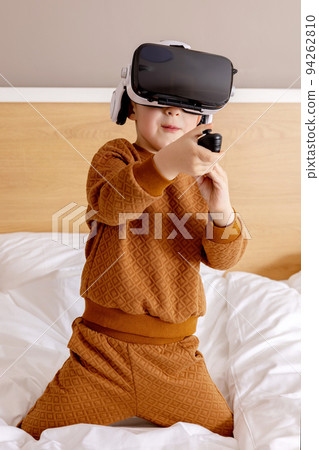 Little adorable boy sitting on bed at home with VR headset and playing interactive video game, exploring virtual reality. Cute child wearing VR glasses. Future, gadgets, technology, education online. 94262810