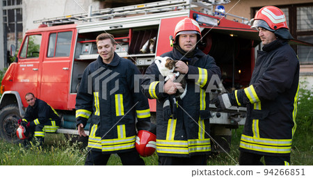 firefighters group in a protective suit and red helmet holds saved cat in his arms. Firefighter in fire fighting operation. firefighters group in a protective suit and red helmet holds saved cat in his arms. Firefighter in fire fighting operation. 94266851