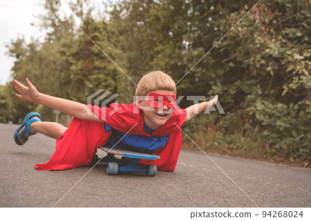 Super hero boy skating on a skateboard outdoors on the road with red glasses and cloak. Kid playing outdoors. Super hero boy skating on a skateboard outdoors on the road with red glasses and cloak. Kid playing outdoors. 94268024