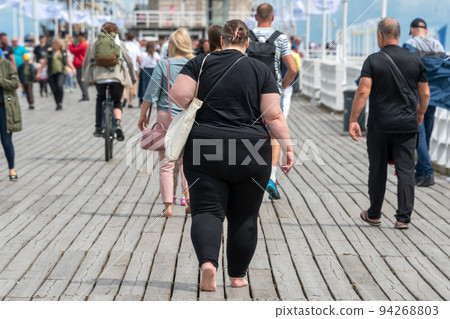Overweight woman walking on the pier among people with normal weight. Barefoot obese woman wearing black. Obesity, losing weight. Overweight woman walking on the pier among people with normal weight. Barefoot obese woman wearing black. Obesity, losing weight. 94268803