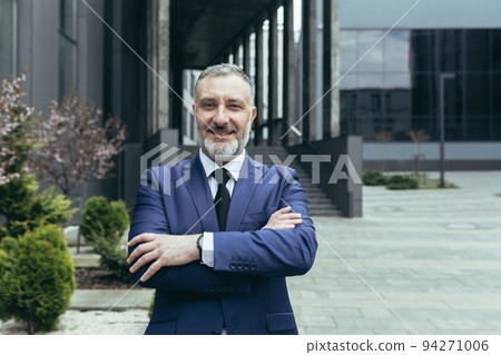 Portrait of a successful senior handsome man, businessman, judge, lawyer. Standing in a suit near the courthouse, office center. Crossed his arms in front, looks at the camera, smiles. 94271006