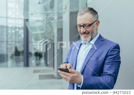 Happy senior handsome gray-haired man, businessman in suit and glasses. Holds the phone in his hands, received good news, reads the message. It stands on the street near a modern building Happy senior handsome gray-haired man, businessman in suit and glasses. Holds the phone in his hands, received good news, reads the message. It stands on the street near a modern building 94271022