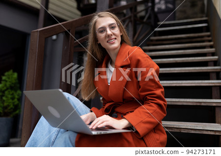 young woman working online on a laptop in an autumn coat sitting on the stairs in the city young woman working online on a laptop in an autumn coat sitting on the stairs in the city 94273171