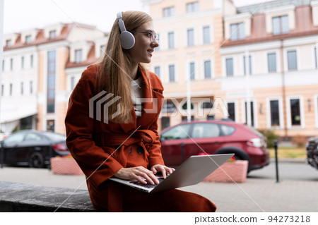 a young girl student in an autumn coat works on a laptop online and listens to music through 94273218