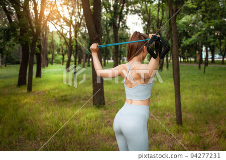 A beautiful young woman performs an exercise outdoor. Grass, park, sports workout. Fitness. An expander. Sports equipment, sunset. A healthy lifestyle, trained muscular body. Rear view, back A beautiful young woman performs an exercise outdoor. Grass, park, sports workout. Fitness. An expander. Sports equipment, sunset. A healthy lifestyle, trained muscular body. Rear view, back 94277231