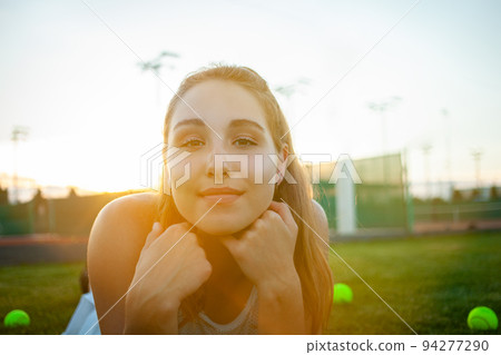 Portrait of a beautiful young woman at sunset. Tennis court, athletic body. Fitness, weight loss, athlete smiles, happy. Active lifestyle in the open air. Sunny glare, enjoyment lighting, background Portrait of a beautiful young woman at sunset. Tennis court, athletic body. Fitness, weight loss, athlete smiles, happy. Active lifestyle in the open air. Sunny glare, enjoyment lighting, background 94277290