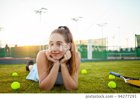 Portrait of a beautiful young woman at sunset. Tennis court, athletic body. Fitness, weight loss, athlete smiles, happy. Active lifestyle in the open air. Sunny glare, enjoyment lying on the grass Portrait of a beautiful young woman at sunset. Tennis court, athletic body. Fitness, weight loss, athlete smiles, happy. Active lifestyle in the open air. Sunny glare, enjoyment lying on the grass 94277291