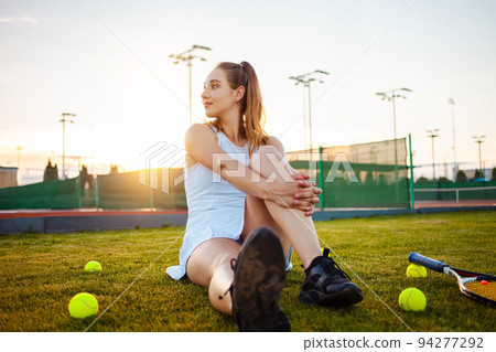 Portrait of a beautiful young woman at sunset. Tennis court, athletic body. Fitness, weight loss, athlete smiles, happy. Active lifestyle in the open air. Sunny glare, enjoyment Rest after training Portrait of a beautiful young woman at sunset. Tennis court, athletic body. Fitness, weight loss, athlete smiles, happy. Active lifestyle in the open air. Sunny glare, enjoyment Rest after training 94277292