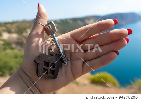 Keys with a keychain in the shape of a house in a female hand against the backdrop of the sea. The concept of buying a house, apartment, real estate, young family, mortgage loan, rental housing. 94278206