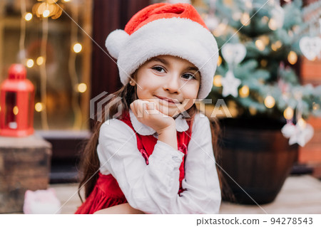 Portrait of emotional smiling cute kid girl in Santa hat and red dress sitting on the porch of a house with decorated Christmas tree. Waiting for Santa and Christmas holidays. Selective focus 94278543