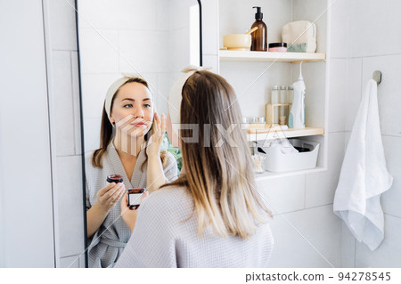 Young woman in bathrobe looking in the mirror and applying facial natural cosmetic clay mask on her face in bathroom. Cosmetic procedures for skin care at home. Beauty self-care. Selective focus. Young woman in bathrobe looking in the mirror and applying facial natural cosmetic clay mask on her face in bathroom. Cosmetic procedures for skin care at home. Beauty self-care. Selective focus. 94278545
