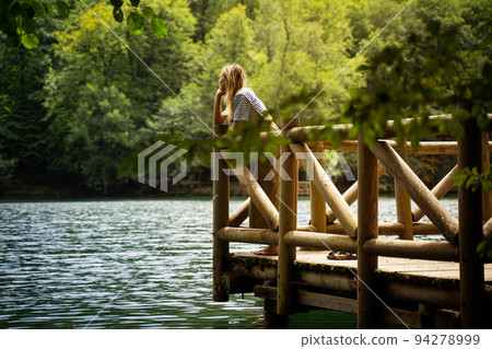 Caucasian blonde woman relaxing on wooden dock by a lake surrounded by green forest in Yedigoller National Park Turkey Caucasian blonde woman relaxing on wooden dock by a lake surrounded by green forest in Yedigoller National Park Turkey 94278999