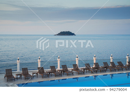 wooden sunlounger chairs near a pool with sea and a small island in the background at sunset 94279004