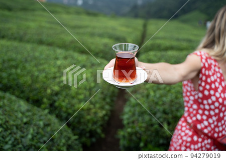 woman in red dress offering a traditional glass of Turkish black tea with rows of tea plantations on terraced hills in the background 94279019