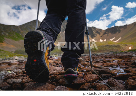 Hiking boots close-up on mountain rocks by the lake with trekking poles, view from below Hiking boots close-up on mountain rocks by the lake with trekking poles, view from below 94282918
