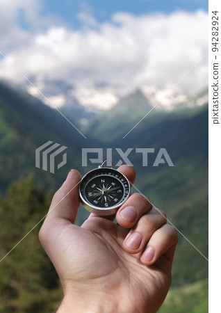 Magnetic compass in the palm of a male hand against the backdrop of a mountain range in the clouds in the summer outdoors, travel, first-person view Magnetic compass in the palm of a male hand against the backdrop of a mountain range in the clouds in the summer outdoors, travel, first-person view 94282924