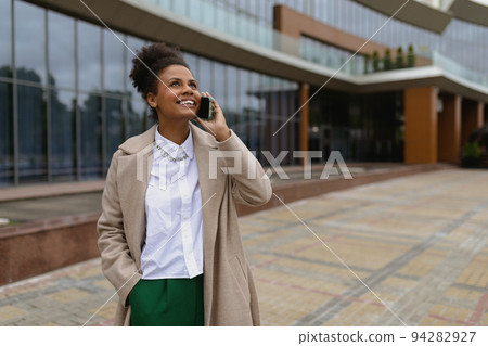 pleased african american young woman talking on mobile phone against glass business center pleased african american young woman talking on mobile phone against glass business center 94282927