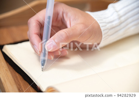 Close-up of female hands writing with gel black pen in leather notepad on wooden table at home 94282929