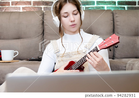 Young caucasian hipster woman is engaged in online ukulele lesson at home, sitting on the floor, on the background of the sofa, in the living room Young caucasian hipster woman is engaged in online ukulele lesson at home, sitting on the floor, on the background of the sofa, in the living room 94282931